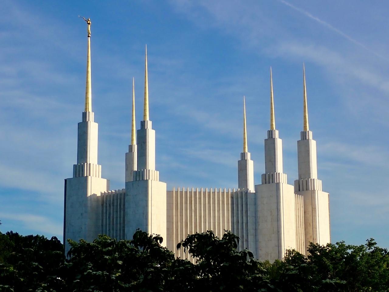 The Washington DC Temple, visible from the treeline, sits majestically as it stretches to the blue sky coated with wispy clouds.